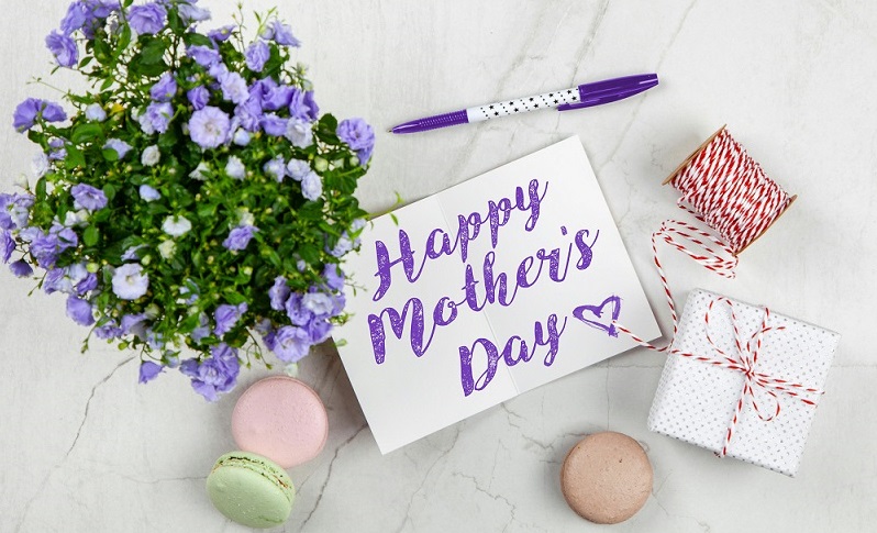 Picture of a bouquet of purple and white flowers with macaroons scattered around the image. A pen, small white gift box and a small sign that reads "Happy Mother's Day" in purple writing are also scattered. The background is white marble.