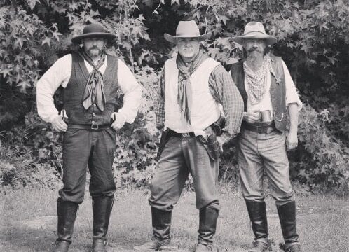 a group of three cowboys in boots, jeans, vests, and cowboy hats stand in a field with trees in the background. the photo is black and white